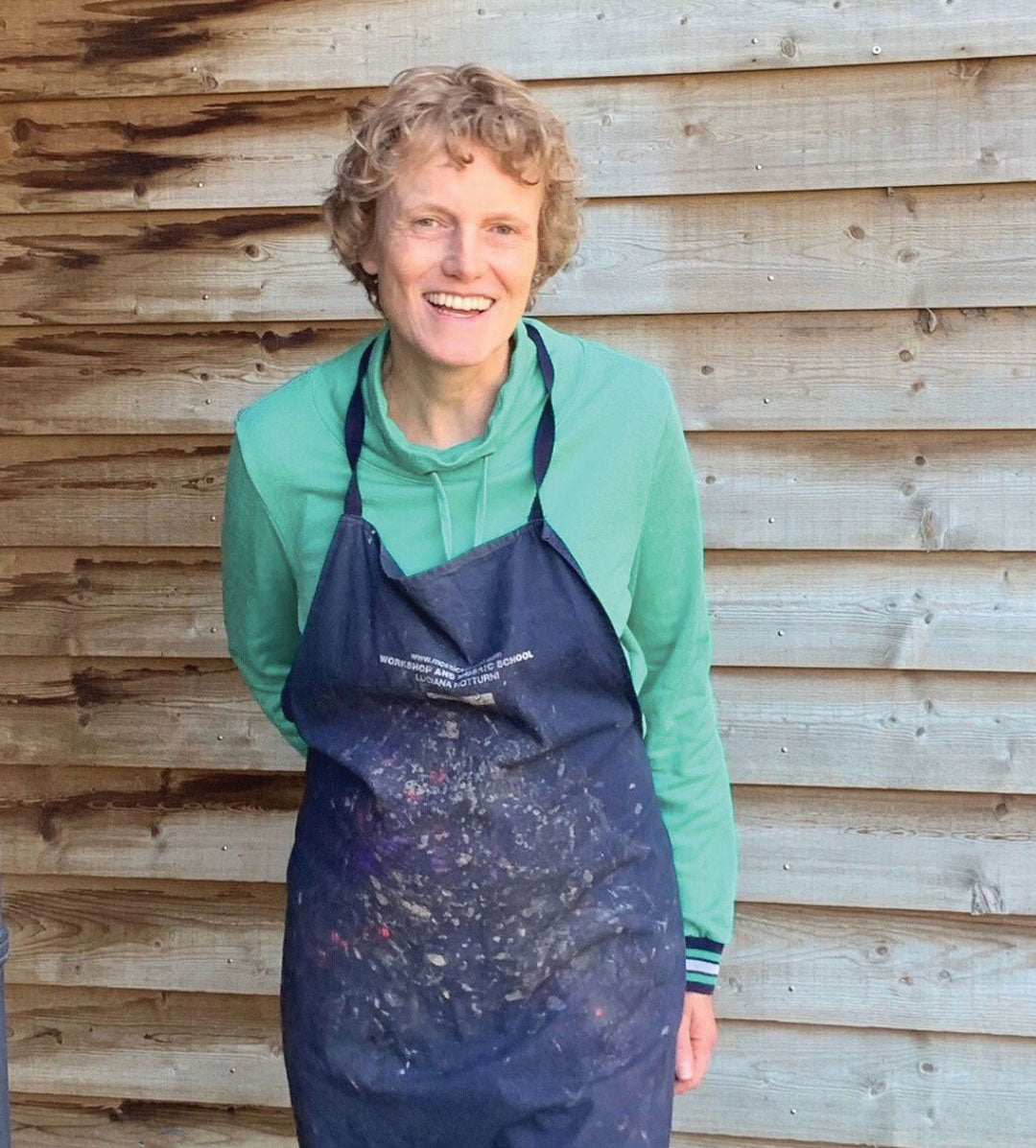 Maartje de Boer smiling in her paint-splattered apron, standing in front of the wooden wall of her studio.
